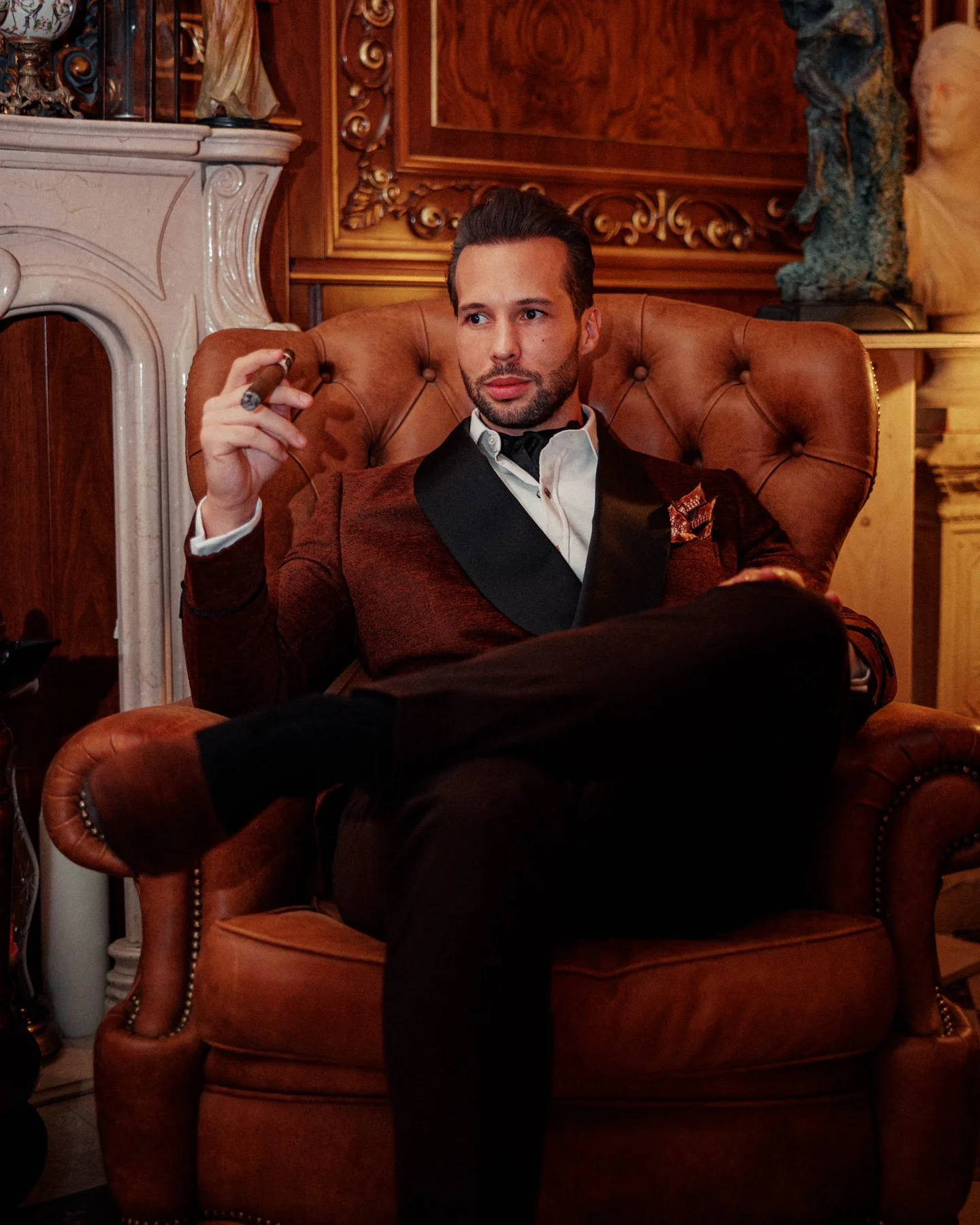 Tristan Tate in a burgundy tuxedo jacket and black bow tie, holding a lit cigar while seated cross-legged in a brown tufted leather armchair, in an opulent room with a marble fireplace and classical statues behind him