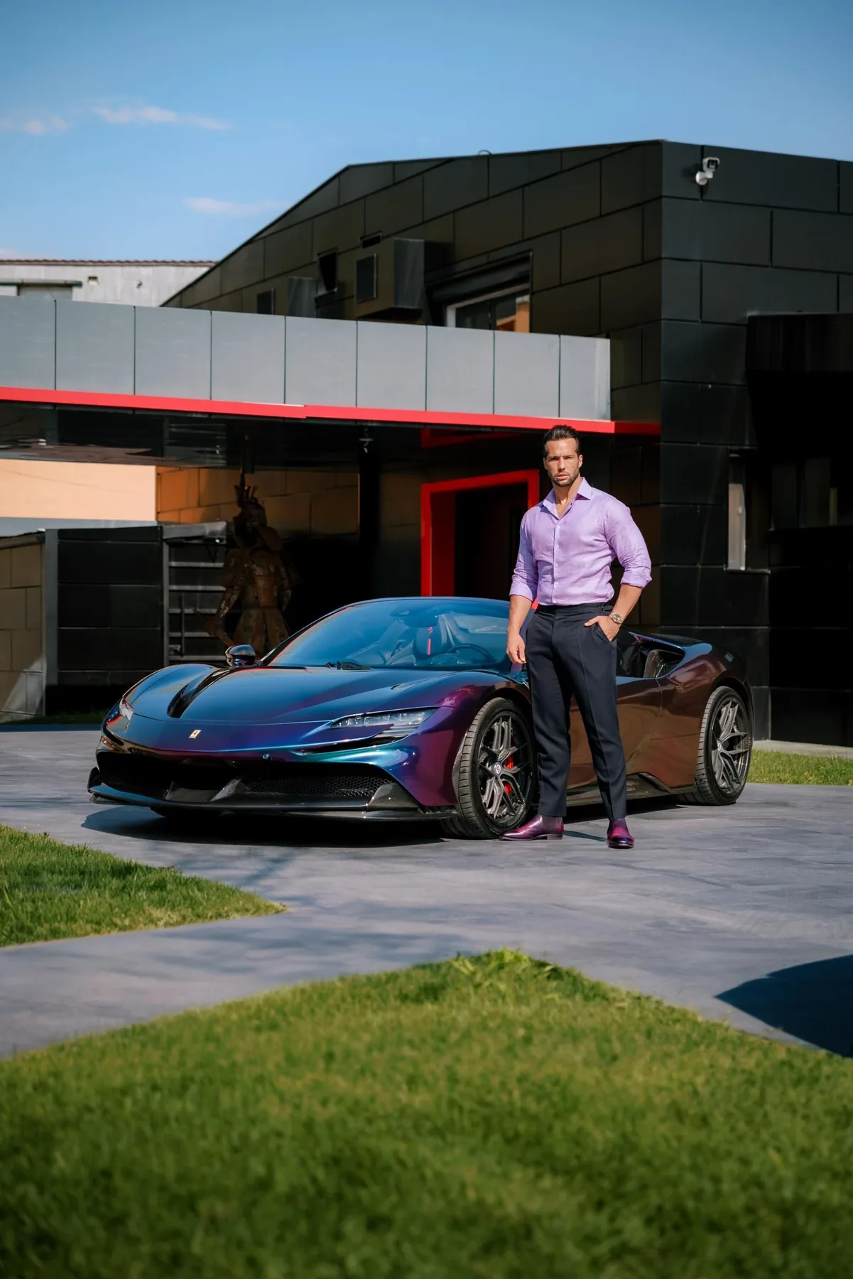 Tristan Tate in a purple linen shirt and navy trousers, hand in pocket, standing beside an iridescent purple-and-green Ferrari SF90 on a paved driveway in front of a modern black-panelled house with red trim