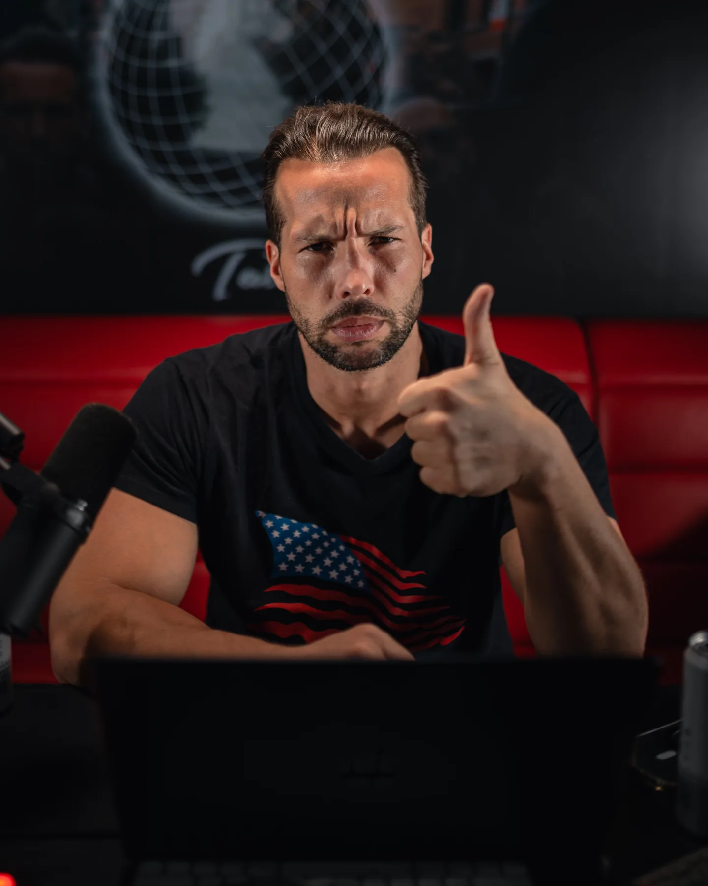 Tristan Tate in a black American-flag t-shirt giving a thumbs up at his podcast desk, with a microphone and laptop in front of him and a red leather couch behind, in a dark studio